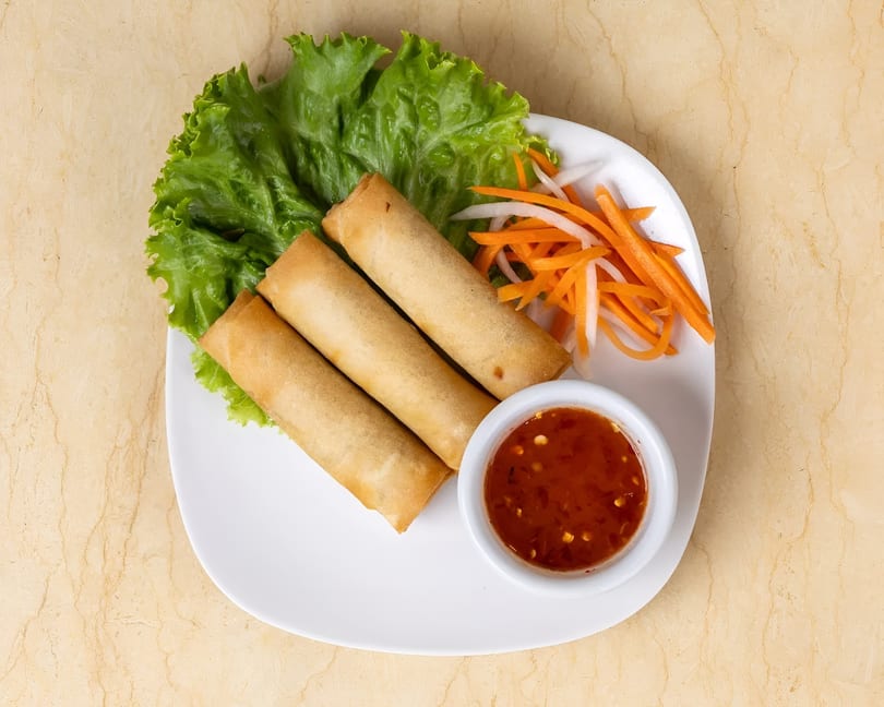 Three golden spring rolls on lettuce with shredded carrots, daikon, and sweet chili sauce.