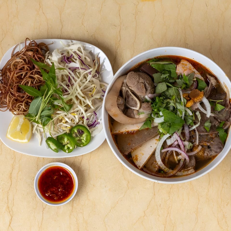 Overhead view of a bowl of Vietnamese soup with meat and vegetables, alongside a plate of garnishes.