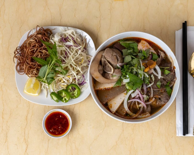 Overhead view of a bowl of Vietnamese soup with meat and vegetables, alongside a plate of garnishes.