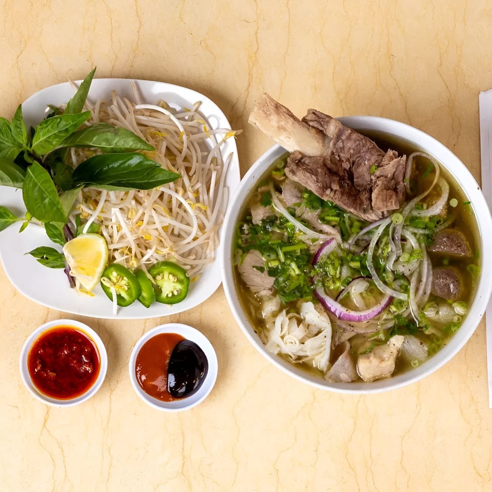 A bowl of pho with a large bone-in beef shank, accompanied by fresh herbs, bean sprouts, and sauces.