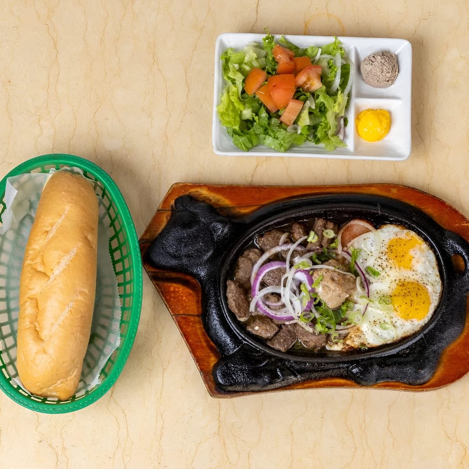 Sizzling beef and eggs with a side salad, pate, mustard, and a baguette in a green basket.