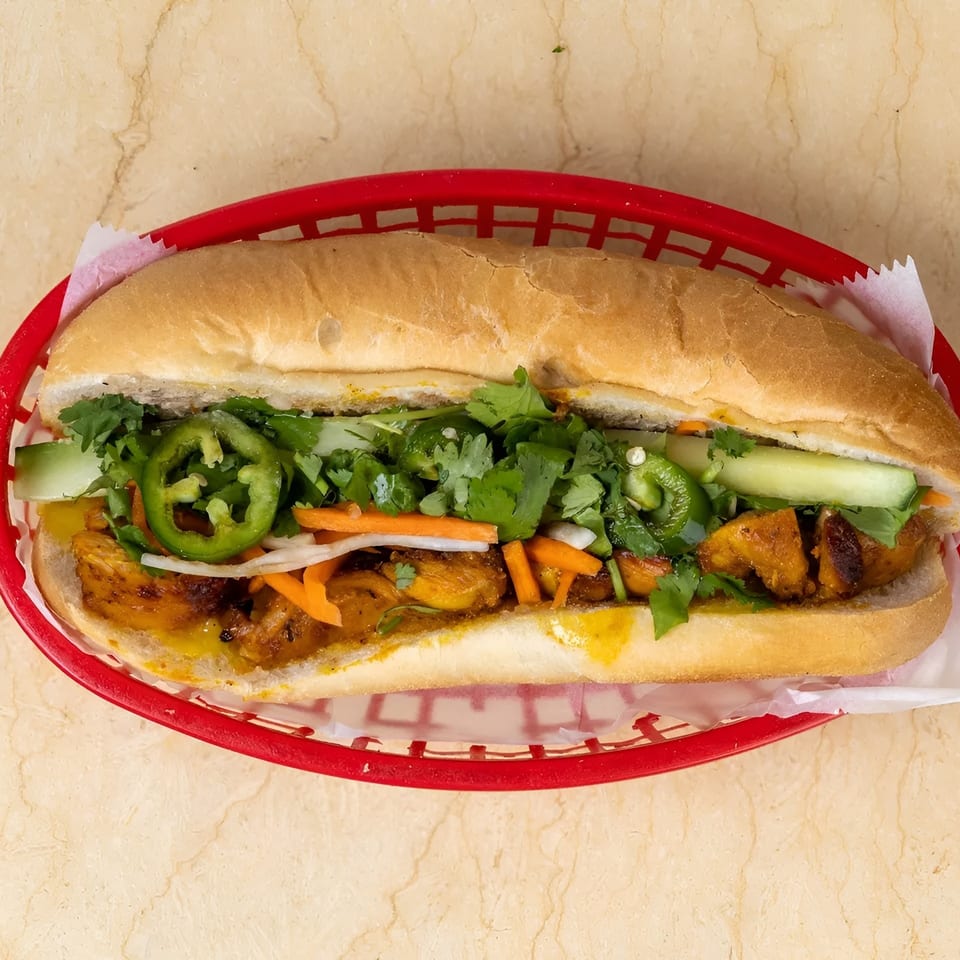 A banh mi sandwich with chicken, cilantro, jalapenos, and pickled vegetables in a red basket.